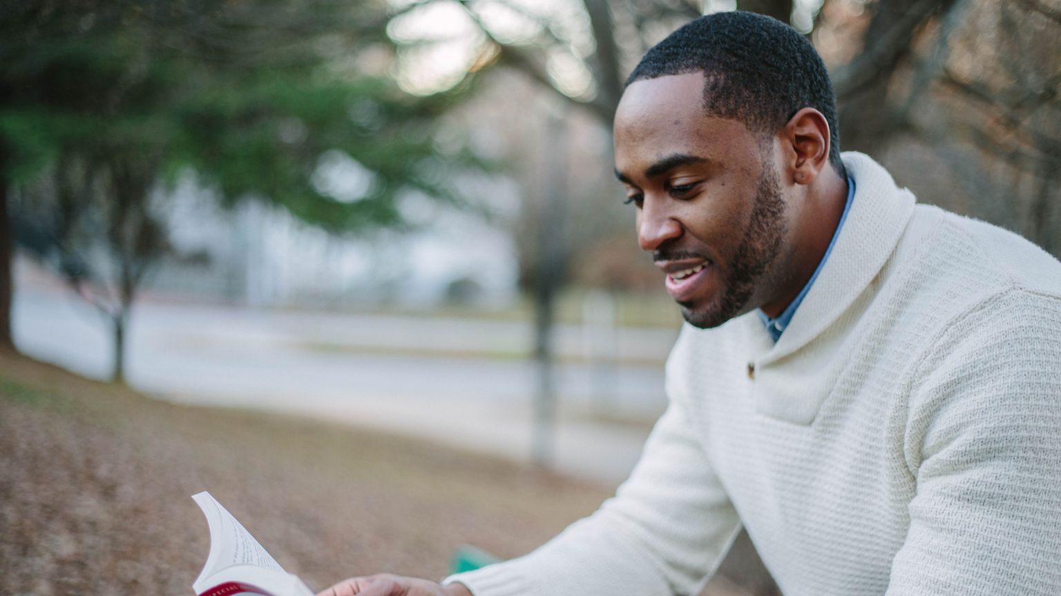 A smiling man in a cream sweater reads a book aloud while sitting outdoors in a park in autumn