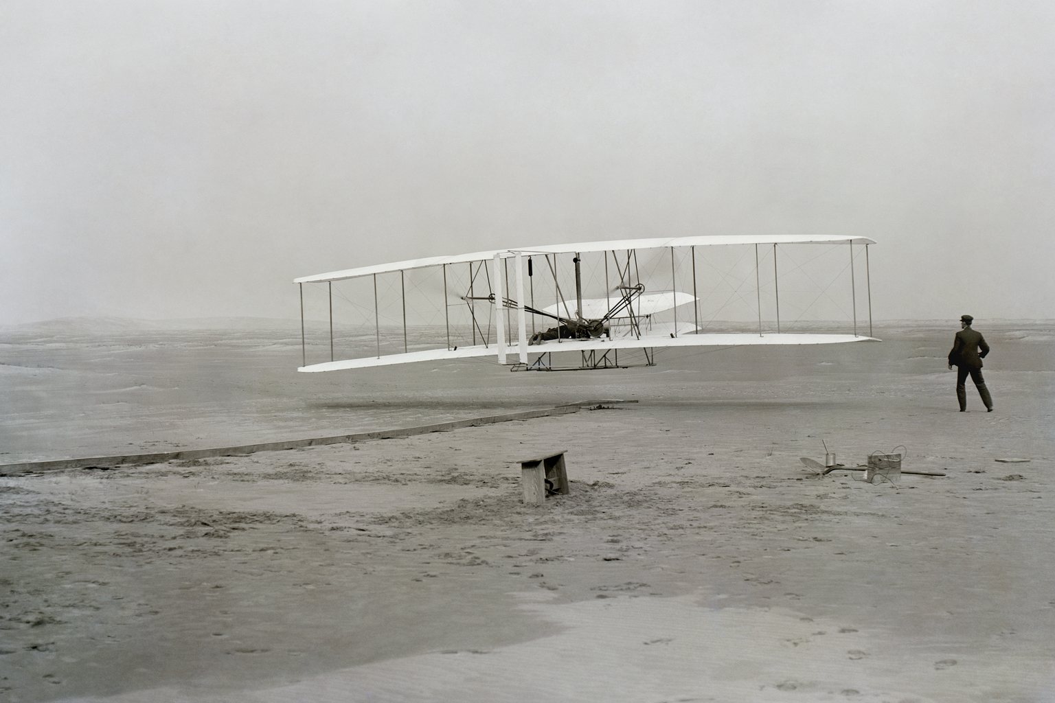 The Wright brothers&rsquo; biplane lifting off a sandy beach at Kitty Hawk in 1903, with Wilbur Wright standing on the sand to the right