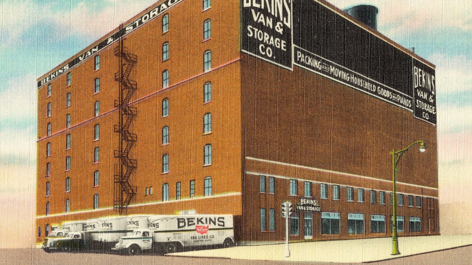 A vintage color postcard of the brick Bekins Van & Storage building in Omaha, with “Packing and moving household goods” painted on the wall and moving vans parked out front