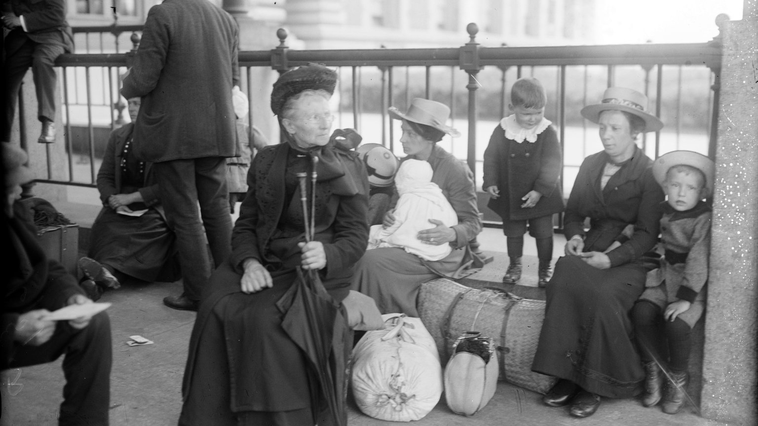 Historic photograph of a Dutch immigrant family with small children and bundled luggage, seated by a railing at Ellis Island