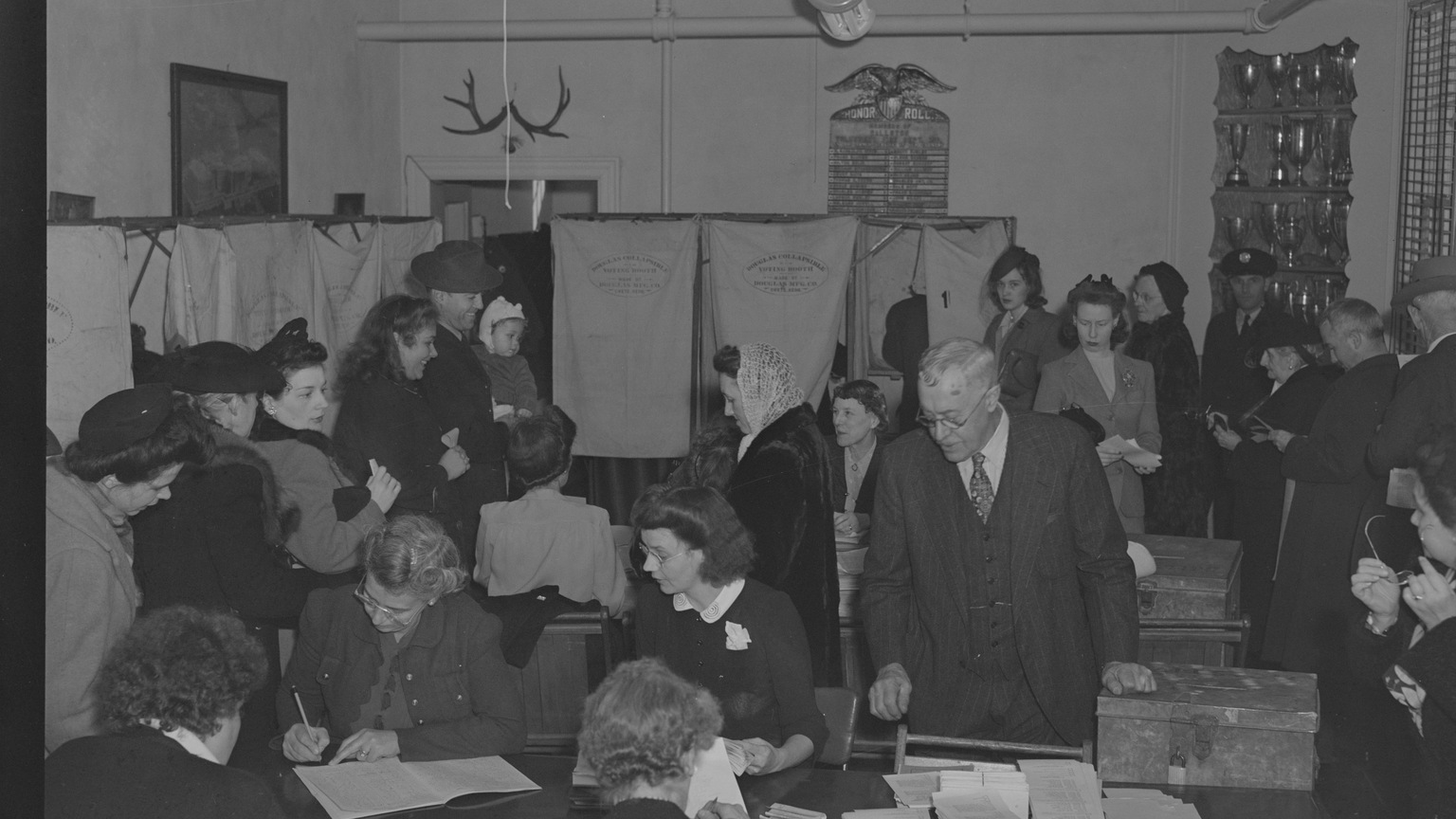 Crowded 1940s polling place with election officials, ballot boxes, and curtained voting booths