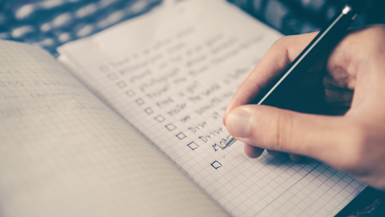 A hand with a pen writing a checklist with empty checkboxes in a grid notebook