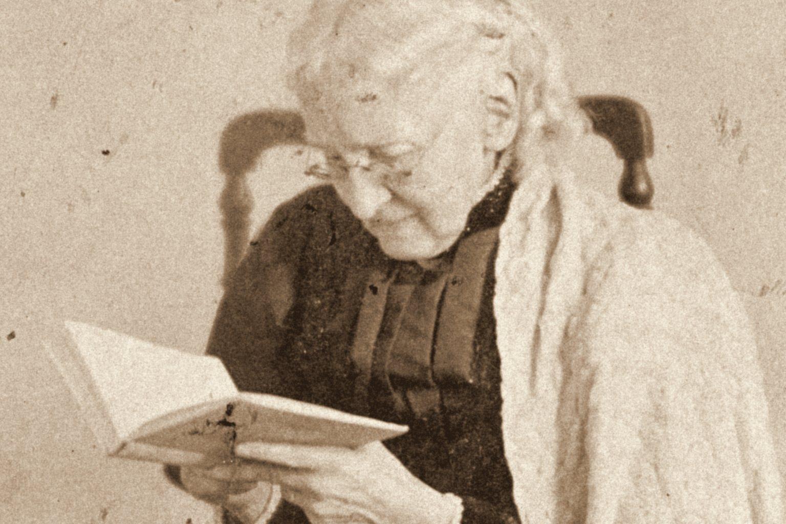 Vintage photograph of an older woman in reading glasses studying an open book, seated in a wooden chair with a shawl