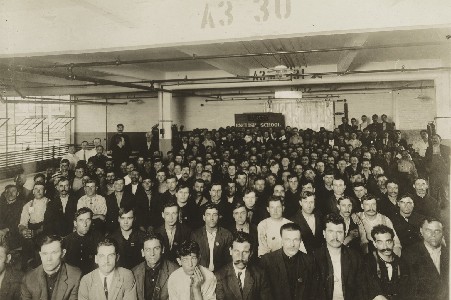A large group of immigrant workers seated in rows in a Ford Motor Company factory room, under a sign that reads English School