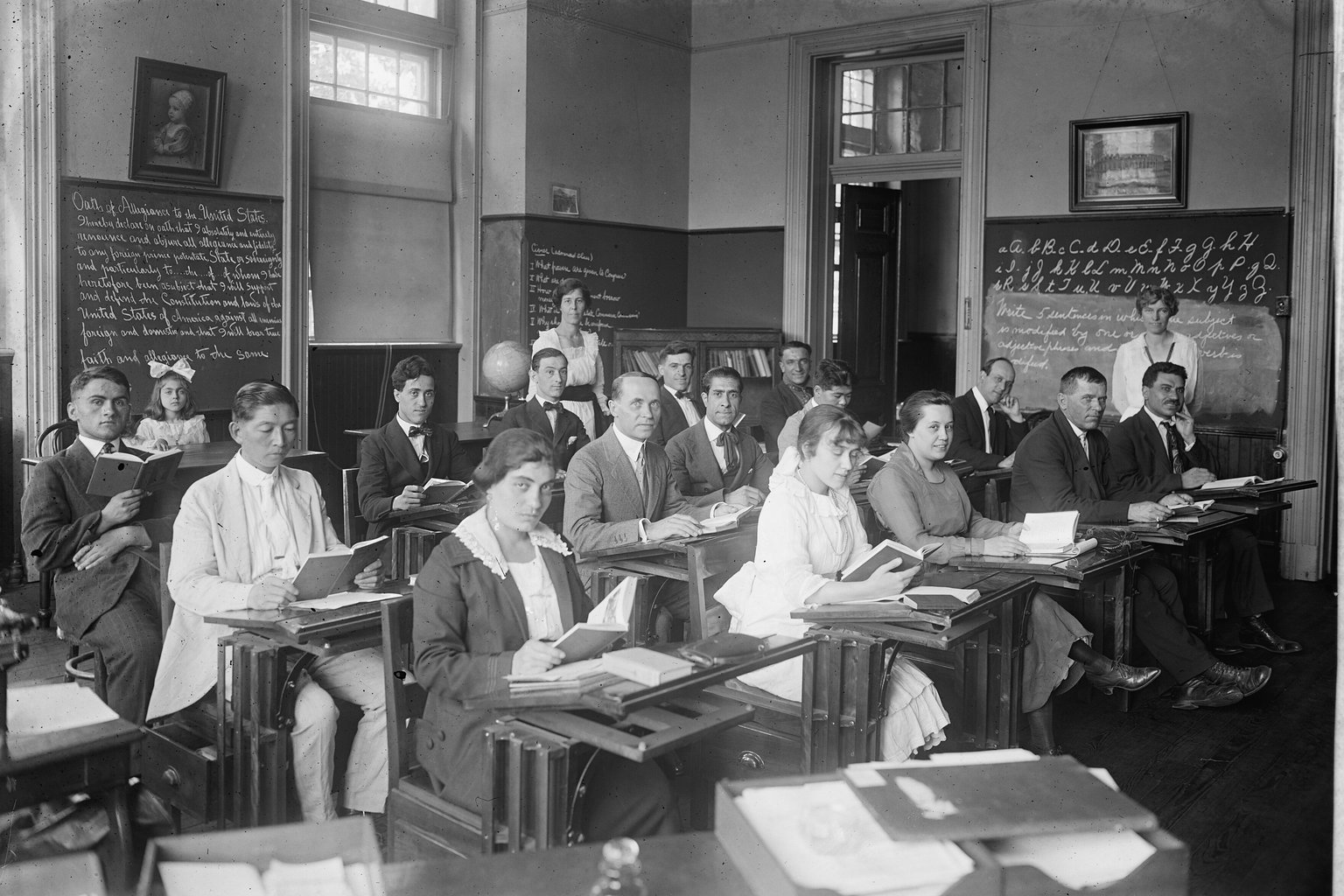 An early 1900s Americanization class of adult students reading at desks, with the Oath of Allegiance written on the blackboard behind them