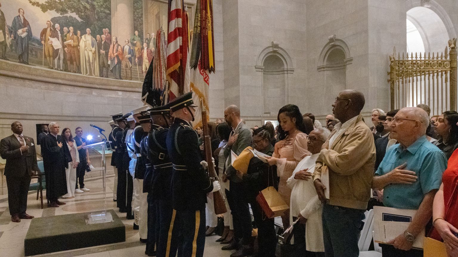 A military color guard presents the flags as new citizens stand with hands on their hearts in the National Archives Rotunda, under a large mural of the Constitution’s framers