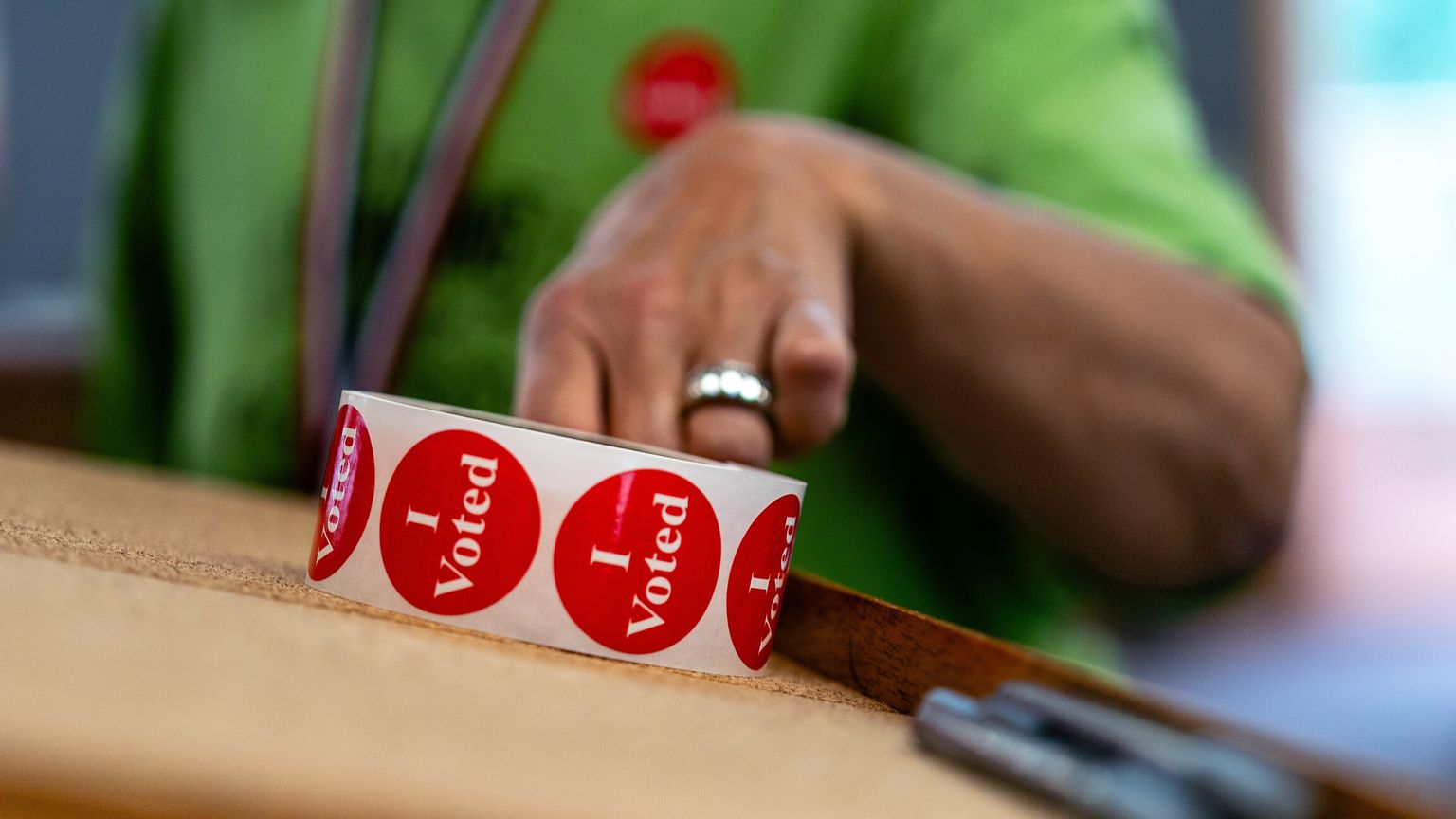 An election judge in a green shirt reaches for a roll of red &ldquo;I Voted&rdquo; stickers on a table at a town hall polling place