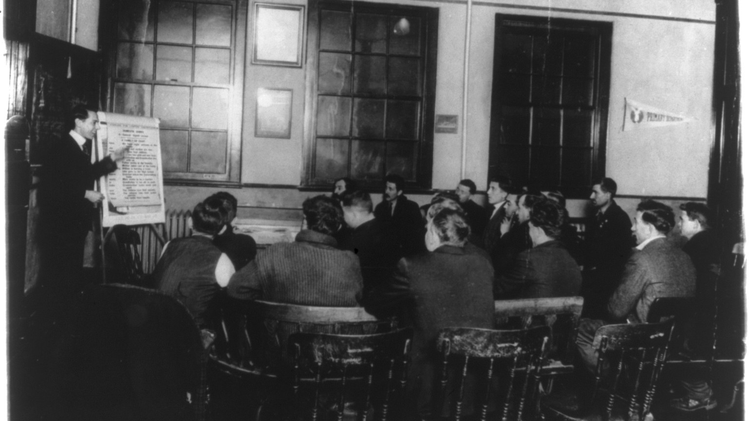Historic photograph of an adult English and citizenship class with an instructor pointing to a lesson chart