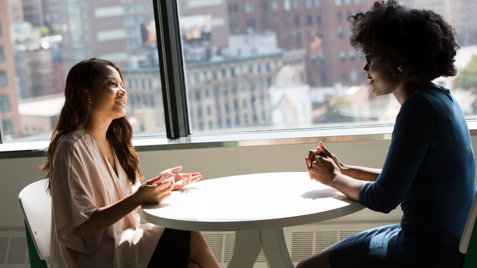 Two women sit across a small white table by a big window, one talking and gesturing while the other listens
