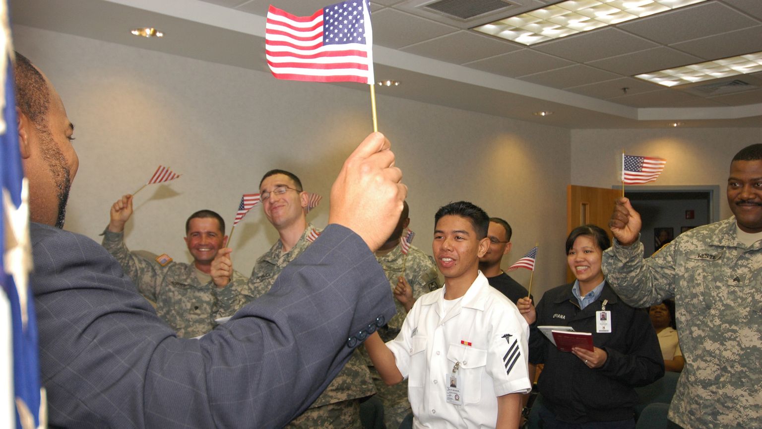 Smiling active-duty sailors and soldiers in uniform wave small American flags after their naturalization ceremony