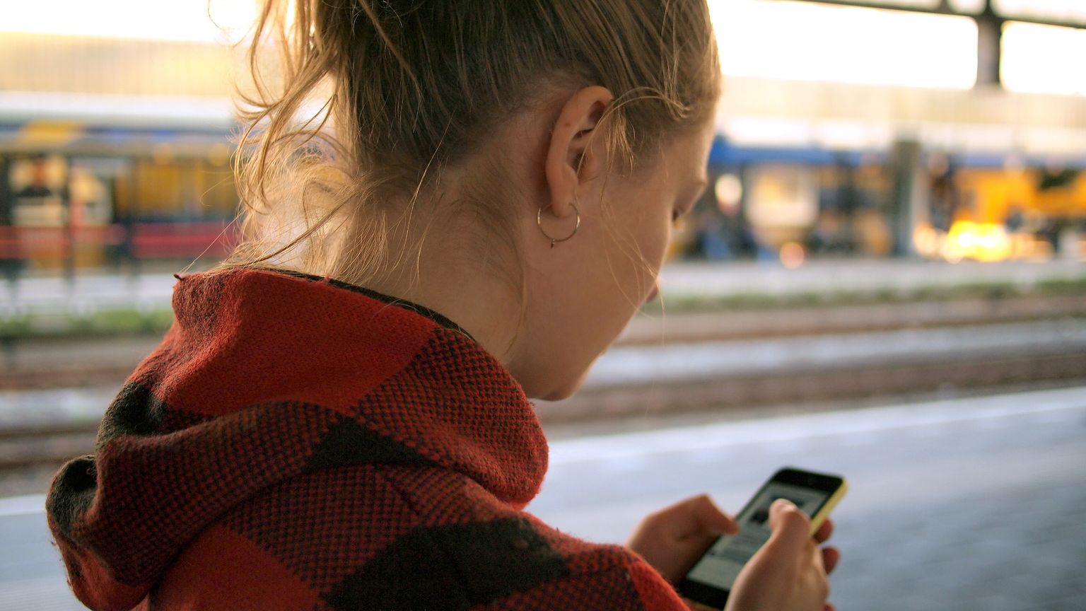 A young woman in a red checked hoodie looks down at her smartphone on a sunny train platform