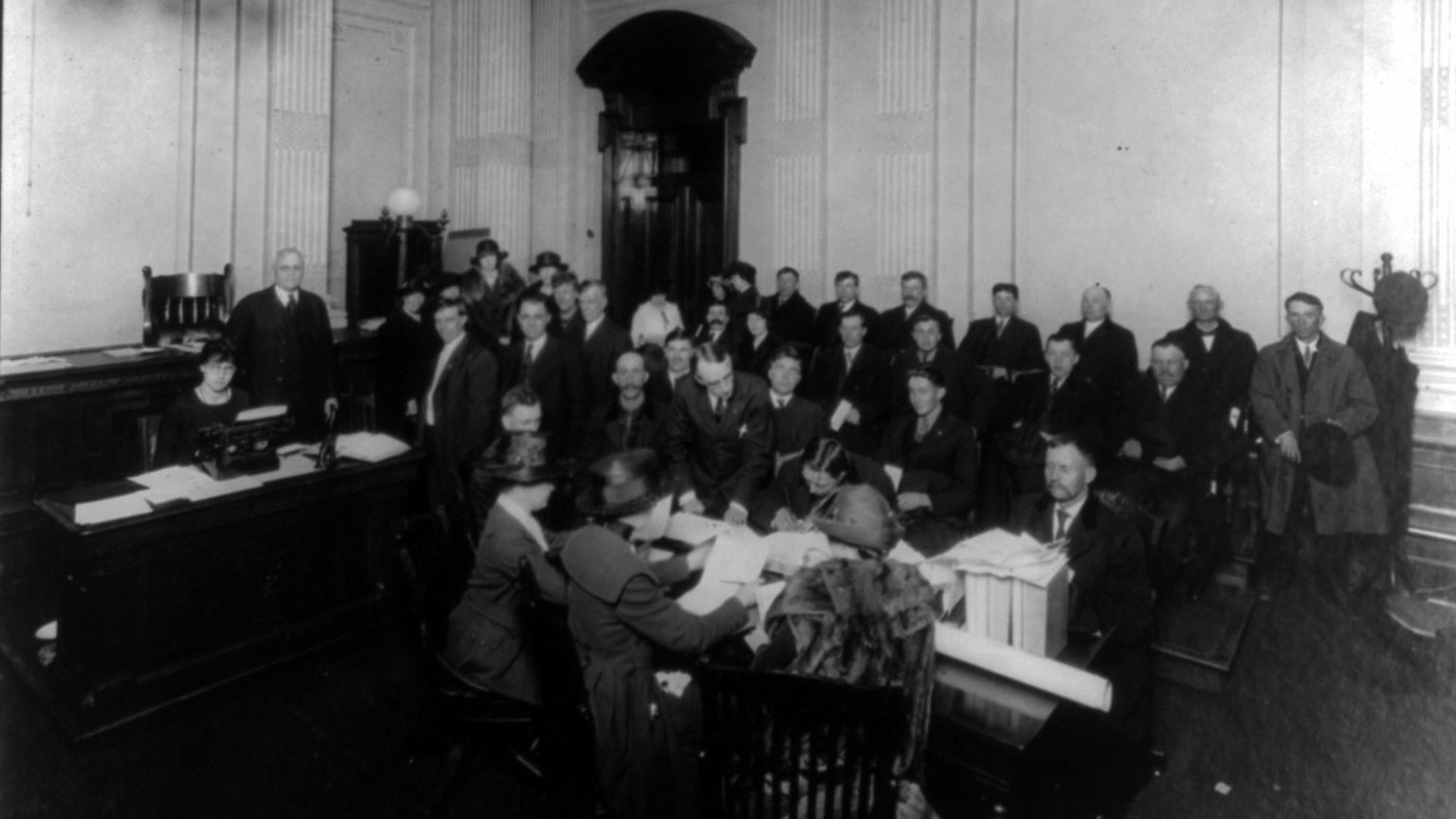 Historic photograph of naturalization applicants seated and standing in a government office while clerks at a desk review their papers