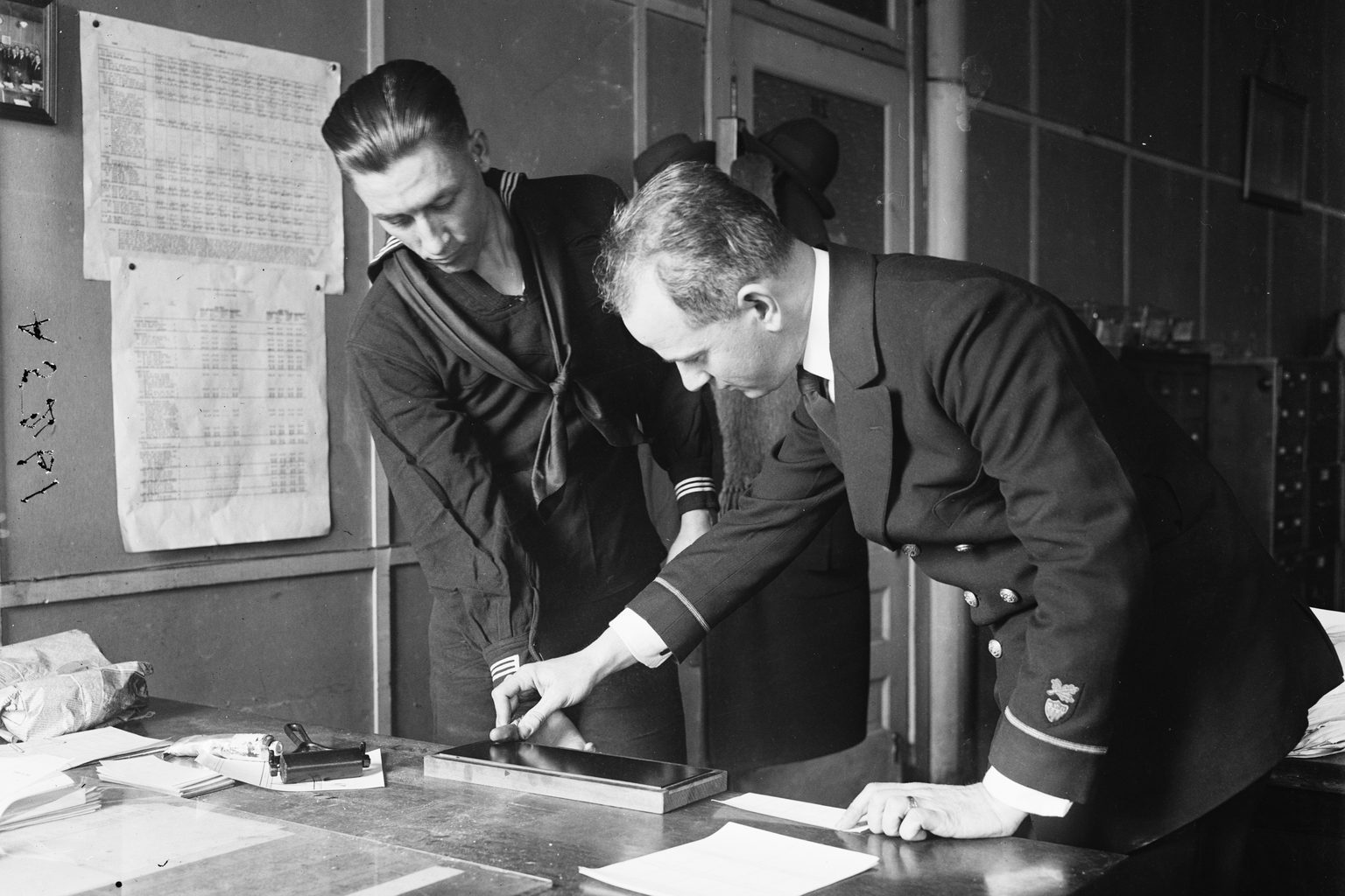 A 1923 photograph of a clerk in a dark uniform guiding a young sailor&rsquo;s finger onto an ink plate to take his fingerprints at a desk