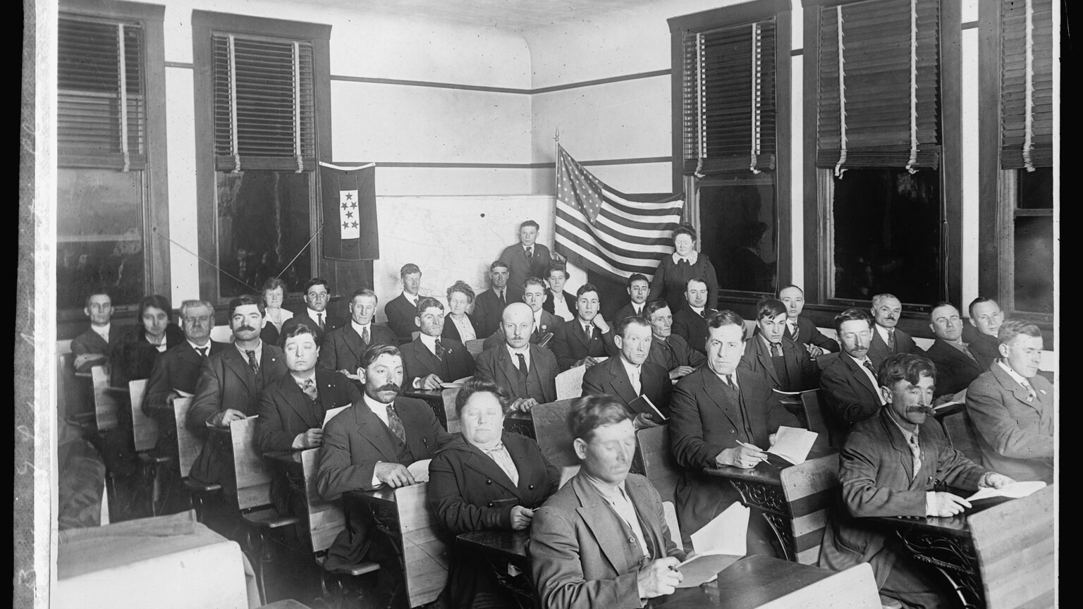 Historic photograph of adults seated in a classroom for a naturalization / citizenship study class with an American flag on the wall