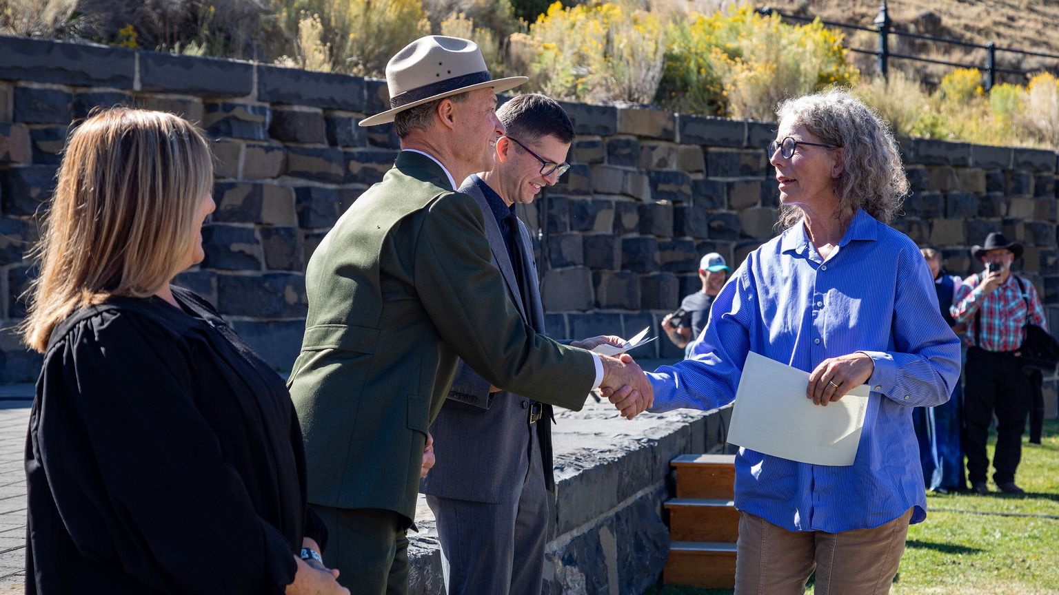 A new citizen in a blue shirt holds her naturalization certificate and shakes hands with a park ranger at an outdoor ceremony