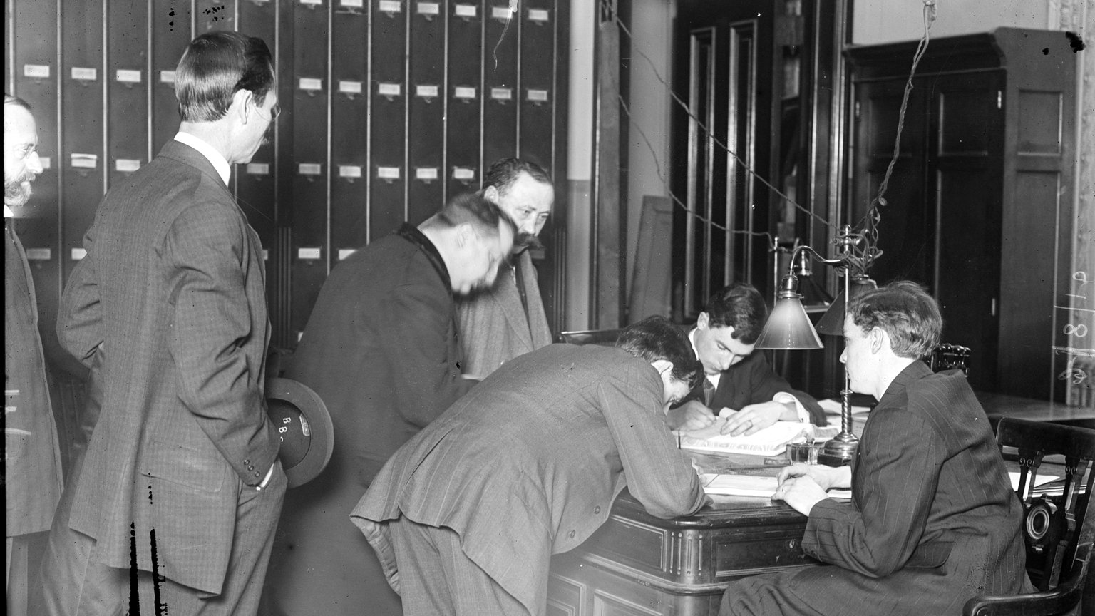 Historic photograph of men in suits leaning over a desk to sign naturalization papers in a judge&rsquo;s chambers, around 1910