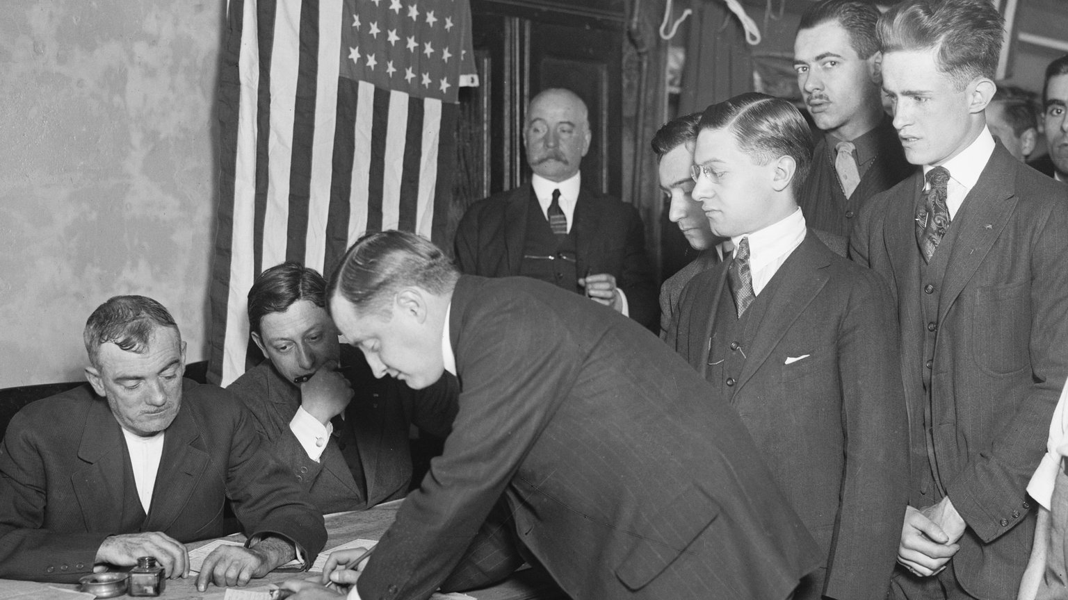 Historic photograph of a young man leaning over a table to sign a draft registration card while other young men wait in line in front of an American flag, New York City, 1917