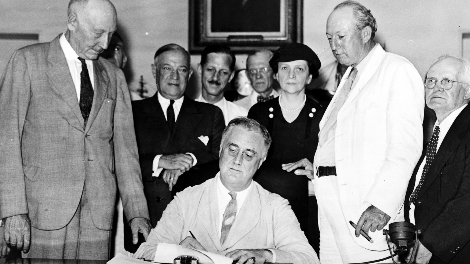 President Franklin D. Roosevelt seated at a desk signing the Social Security Act in 1935, with Labor Secretary Frances Perkins and members of Congress standing behind him