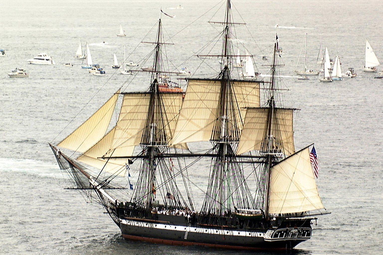 The wooden frigate USS Constitution, Old Ironsides, under full sail on gray water with small boats in the distance