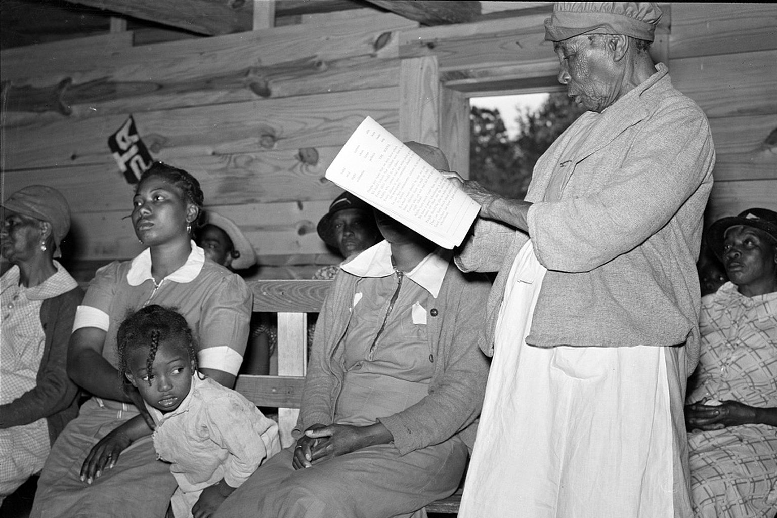An 82-year-old woman stands and reads her lesson aloud from a sheet of paper in an adult class in Gee&rsquo;s Bend, Alabama, in May 1939, while other students sit on benches and listen
