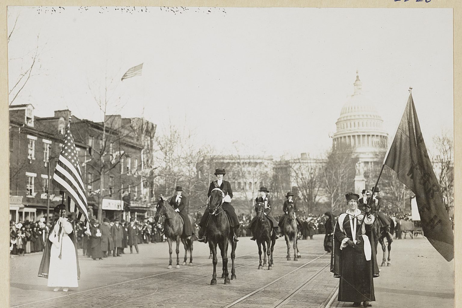 Historic photograph of a 1913 women’s suffrage parade on Pennsylvania Avenue in Washington, D.C., with marchers, a flag, and the U.S. Capitol in the background