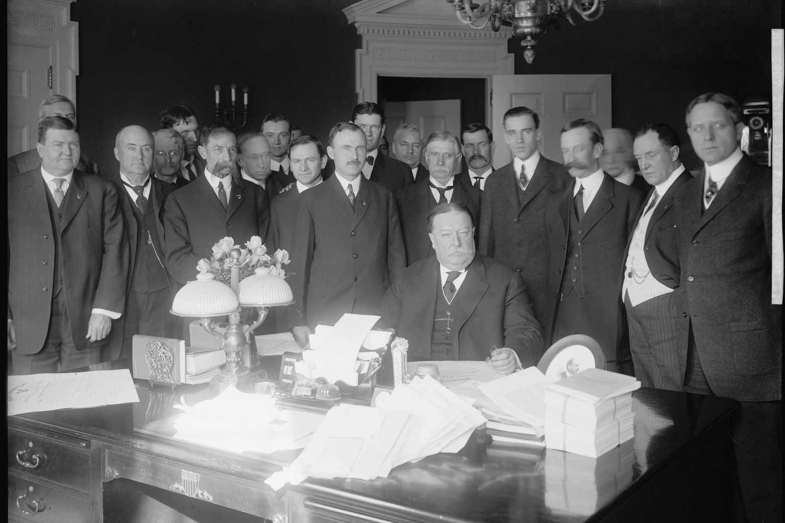 President William Howard Taft seated at his desk signing a bill in 1912, with a group of men standing behind him